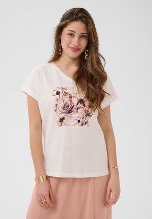 Ung kvinde med langt brunt hår iført en hvid T-shirt med pink blomstertrykk, pink nederdel, gyldne øreringe og armbånd, smilende blidt.