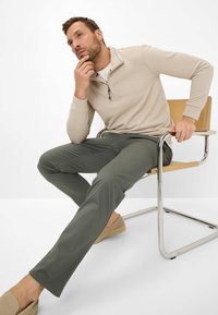 Light beige zip-up sweatshirt, olive green pants, and tan slip-on shoes. Model sits on a chrome chair, hand on chin, with neutral background.