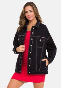 Schwarze Jeansjacke mit wei�ßen Nähten, Fronttaschen und silbernen Knöpfen, getragen über einem figurbetonten roten Kleid mit Knopfverschluss.