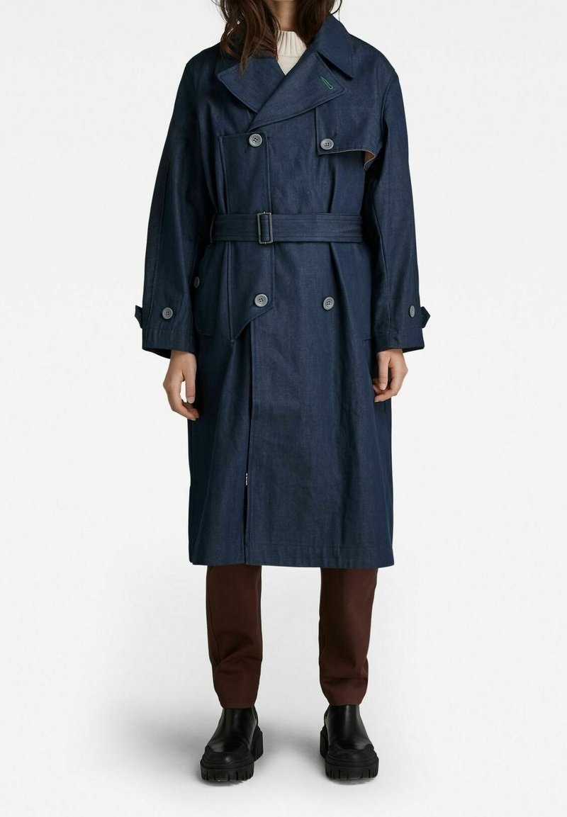 Personne portant un long trench-coat bleu foncé ceinturé, un pantalon marron, des bottes noires épaisses et un top blanc, debout devant un fond uni.