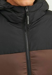 Pehme kinnas, millel on must ülaosa ja pruun alumine osa, ees on lukk ja logo tõmblukk sulgemiseks.