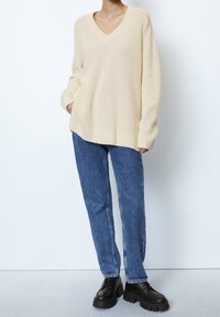 Pull beige oversize à col en V en maille texturée, associé à un jean droit bleu et à des chaussures à plateforme noires à lacets.