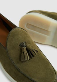 Olijfgroene suède loafers met een ronde neus, leren kwastdetail en een contrasterende witte rubberen zool met een gestructureerde onderkant.