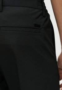 Κοντινό πλάνο στα μαύρα παντελόνια ενός ατόμου που δείχνει μια πίσω τσέπη με μια μικρή ορθογώνια μαύρη ετικέτα κοντά στη ραφή της τσέπης.