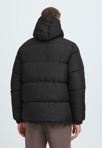 Giacca puffa nera con cappuccio, caratterizzata da sezioni trapuntate, polsini elasticizzati e una vestibilità comoda. Texture liscia, design minimalista.