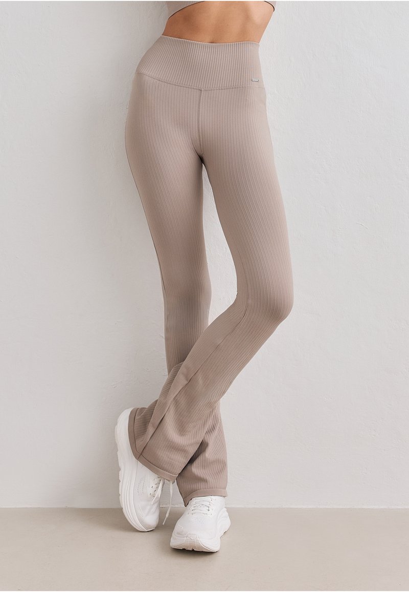 Leggings évasés côtelés beige taille haute, portés avec des baskets blanches, vus de la taille aux pieds sur un mur uni.