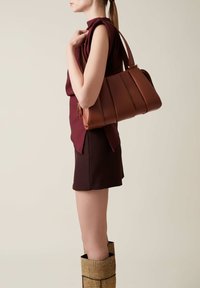 Brown leather handbag with pleated design and gold hardware, worn on arm. Model is dressed in a burgundy top and shorts, with patterned gold boots.