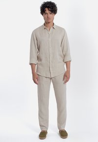 Lichtbeige linnen overhemd en broek met knopen aan de voorkant, rechte snit, opgerolde mouwen en een textuur stof. Gecombineerd met groene instapschoenen.