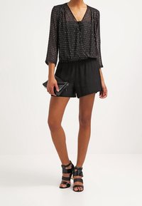 Blusa negra transparente con rayas verticales plateadas, combinada con pantalones cortos negros de cintura alta. Los accesorios incluyen un bolso negro de mano y sandalias negras con tiras.