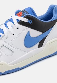 Bílý kožený teniska s modrými a černými akcenty, výrazným modrým logem Nike, texturovanou podrážkou a bílými tkaničkami.