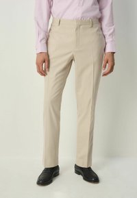 Man i beige kostymbyxor, svarta skor och en ljusrosa långärmad skjorta med knappar, som står mot en neutral bakgrund.