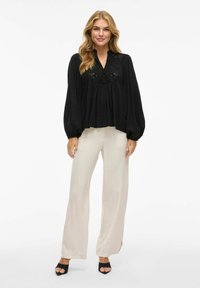 Blusa nera con maniche voluminose e dettagli in pizzo, abbinata a pantaloni cream a gamba larga e tacchi aperti neri.
