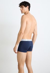 Jeune homme debout torse nu, portant un boxer bleu marine avec une ceinture blanche marquée "GANT", de profil sur un fond uni.