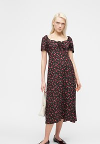 Νέα γυναίκα με ξανθά μαλλιά, ντυμένη με μαύρο μίντι φόρεμα με κόκκινο λουλουδάτο σχέδιο, κρατώντας λευκή τσάντα και φορώντας μαύρα φλατ παπούτσια.