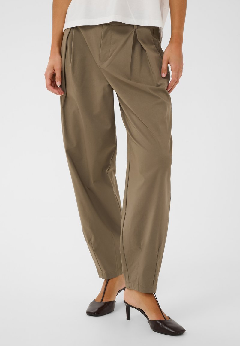 Femme portant un pantalon ample vert olive et des chaussures à talons hauts noires avec de fines brides, debout devant un fond clair uni.