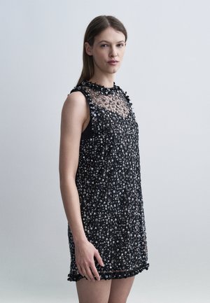 Woman standing sideways wearing sleeveless black dress with sheer upper chest and floral sequin embellishments against plain gray background.