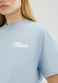 Lichtblauwe t-shirt met een ronde hals, korte mouwen en een witte geborduurde "Ellesse" logo aan de voorkant. Smooth katoen stof textuur.