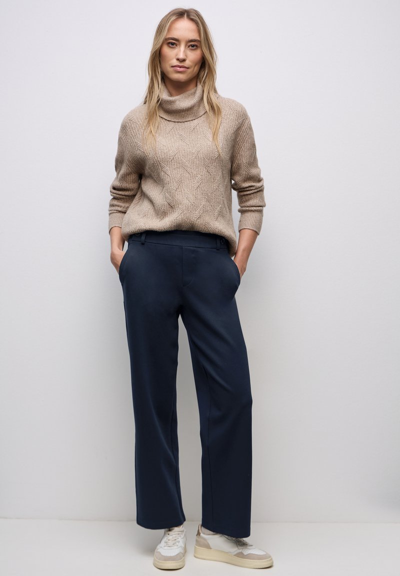 Beżowy sweter z dzianiny z wysokim kołnierzem i teksturowanym wzorem, zestawiony z granatowymi spodniami o szerokich nogawkach i białymi sneakersami. Prosty i nowoczesny design.