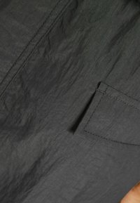 Dunkelgrauer Stoff mit zerknitterter Textur, versehen mit einer kleinen Tasche und kontrastierenden Nähten. Glatte Oberfläche mit dezentem Glanz.