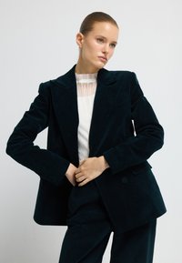 Žena s hladce začenutými vlasy, oblečená v tmavě tyrkysovém sametovém obleku a bílé plisé halence, pózuje na jednobarevném světlém pozadí.