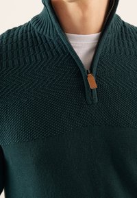 Donkergroene trui met ritskraag, voorzien van textuurrijke gebreide patronen, een glad lichaam en een leren ritslip. Contrasterende patronen aan de bovenkant.