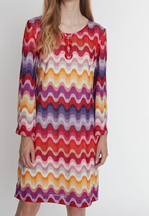 Kvinde iført en langærmet kjole med flerfarvede bølgede striber i rød, orange, lilla og hvid samt tre røde rundede knapper ved halsudskæringen.
