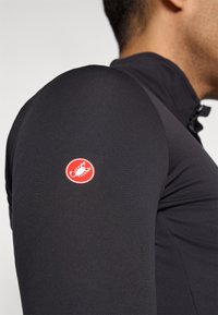 Le maillot de cyclisme à manches longues noir présente un design ajusté, une texture lisse et un logo rouge bien visible sur le bras supérieur. Ouverture zippée au niveau du cou.