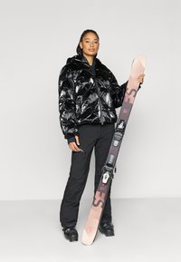 Μαύρο φουσκωτό μπουφάν με σχέδιο καπιτονέ, μαύρο παντελόνι σκι και μαύρα μποτάκια σκι. Κρατάει ένα ανοιχτόχρωμο σκι με δέστρες και σήμανση.