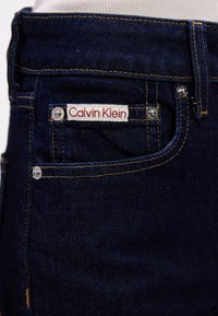 Ciemnoniebieskie dżinsy z białą i czerwoną metką Calvin Klein na tylnej kieszeni, z widocznymi żółtymi szwami i metalowymi nitami.
