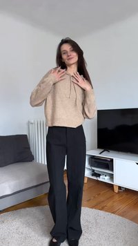 Bézs kötött pulóver kapucnival, fekete, bőszárú nadrággal párosítva. Simább textúrájú, enyhén fénylik; világos szőnyegen áll.