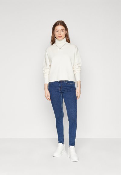 Suéter blanco de cuello alto cortado con mangas largas, combinado con jeans ajustados azules y zapatillas blancas. Texturas simples y suaves evidentes.