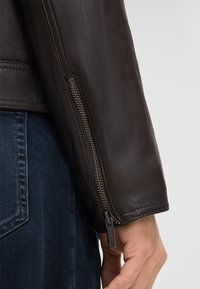 Ärmel einer braunen Lederjacke mit strukturiertem Finish, ausgestattet mit einem metallischen Reißverschluss-Detail am Bündchen. Dunkler Jeanshintergrund sichtbar.