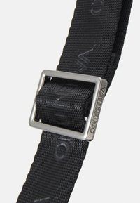 Black fabric belt with a silver rectangular metal buckle. The belt features a raised logo pattern throughout its surface for texture.