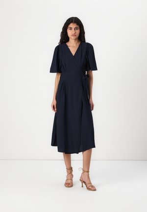 Robe enroulée bleu marine avec des manches bouffantes courtes, un détail à nouer à la taille et une silhouette évasée fluide. Tissu lisse avec une longueur mi-mollet.