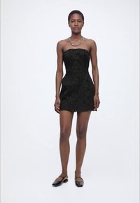 Γυναίκα με μαύρο μίνι φόρεμα δαντέλας χωρίς τιράντες, μαύρα επίπεδα παπούτσια και αλυσίδα στον λαιμό, όρθια μπροστά σε ένα απλό λευκό φόντο.