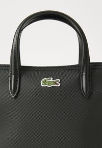 Sac à main noir texturé avec des poignées en cuir brillant. Présente un petit logo crocodile vert à l'avant, positionné sur la gauche.