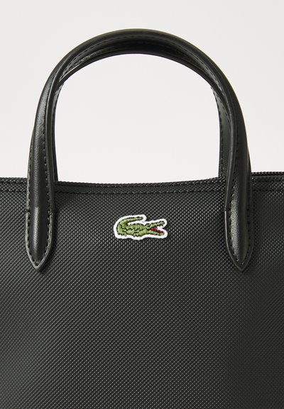 Sac à main noir texturé avec des poignées en cuir brillant. Présente un petit logo crocodile vert à l'avant, positionné sur la gauche.