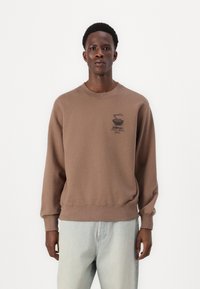 Felpa marrone con scollatura rotonda, polsini e orlo a coste. Presenta un design grafico nero sul petto. Abbinata a jeans azzurri chiari.