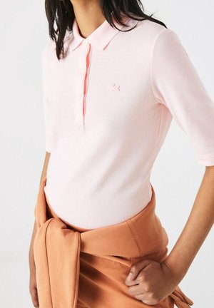 Vrouw draagt lichtroze poloshirt met korte mouwen, kraag en knopen, houdt een beige jasje vast dat rond haar taille is gebonden, tegen een witte achtergrond.