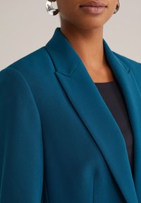 Detailný záber osoby v tehlovej blejzri, čiernom topi a strieborných kruhových náušniciach na svetlom neutrálnom pozadí.