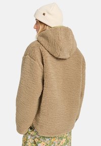 Polerka z polaru w kolorze tan, z teksturowaną powierzchnią i kapturem. Noszona z jasno kolorowym beanie i spodniami w kwiatowy wzór.
