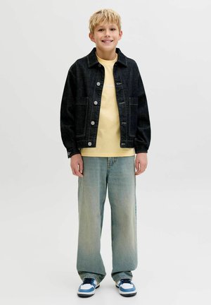 Donkere blauwe spijkerjas met zilverkleurige knopen, gedragen over een lichtgeel shirt. Lichtblauwe wijde spijkerbroek en witte sneakers met blauwe accenten.