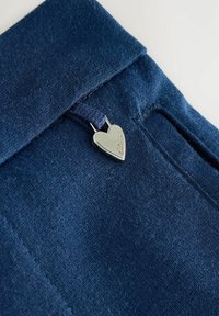 Donkerblauwe stof met een zachte textuur, voorzien van een hartvormige zilveren hardwareaccent dat aan een lus is bevestigd. Het ontwerp bevat een gevouwen tailleband.