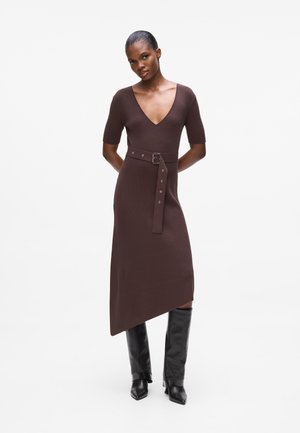 Brown ribbed knit dress with a deep V-neck, short sleeves, side belt with a metallic buckle, and an asymmetrical hem. Worn with tall black boots.