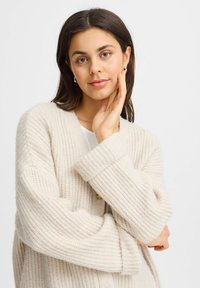 Cardigan beige in maglia con un motivo texturizzato, vestibilità oversize e maniche ampie. Presenta un collo a V e un dettaglio con bottoni.