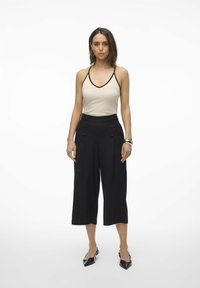 Top beige à bretelles avec des bordures noires, associé à un pantalon large noir taille haute à double boutons, et des chaussures noires à bout pointu.