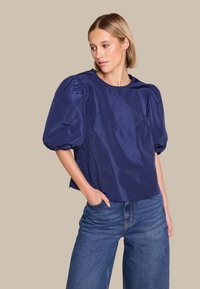 Mørkeblå, lett, satenglignende topp med puffede ermer, rund hals og løs passform, kombinert med casual denimjeans.