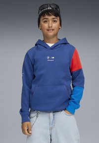 Felpa blu con una manica rossa e blu, una grande tasca frontale e un logo sul petto. Indossata con pantaloni corti in denim chiaro e un accessorio a catena.