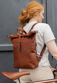 Brązowy skórzany plecak z rolowanym zapięciem, regulowanymi paskami i metalowym zapięciem. Gładka struktura i minimalistyczny styl.