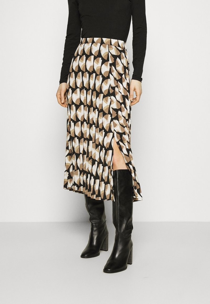 Femme portant un haut noir à manches longues, une jupe midi à motif géométrique avec une fente latérale, et des bottes montantes noires à talons.